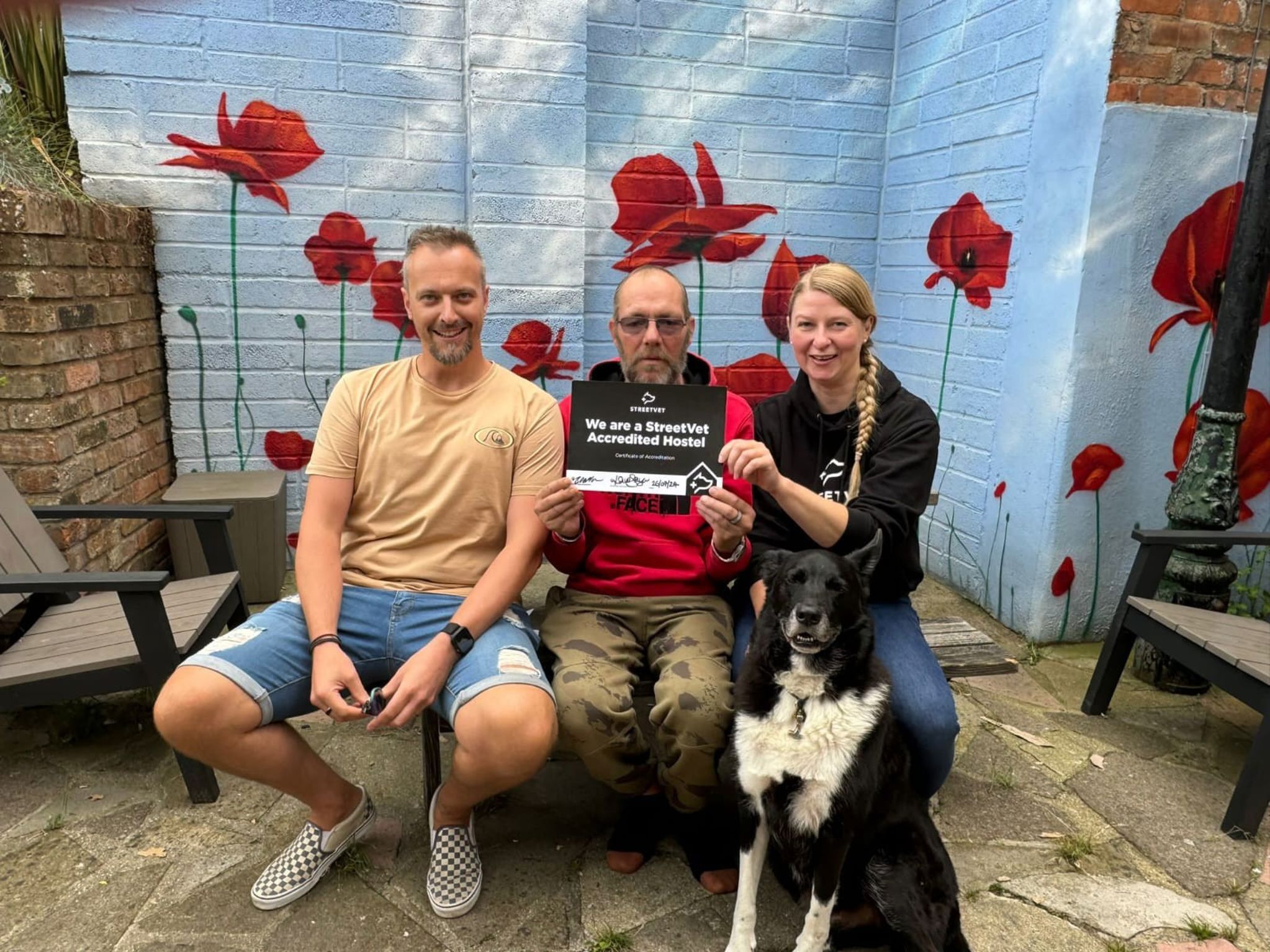 Three people sitting with a black-and-white dog, holding a StreetVet Accredited Hostel certificate in front of a wall painted with red poppies.