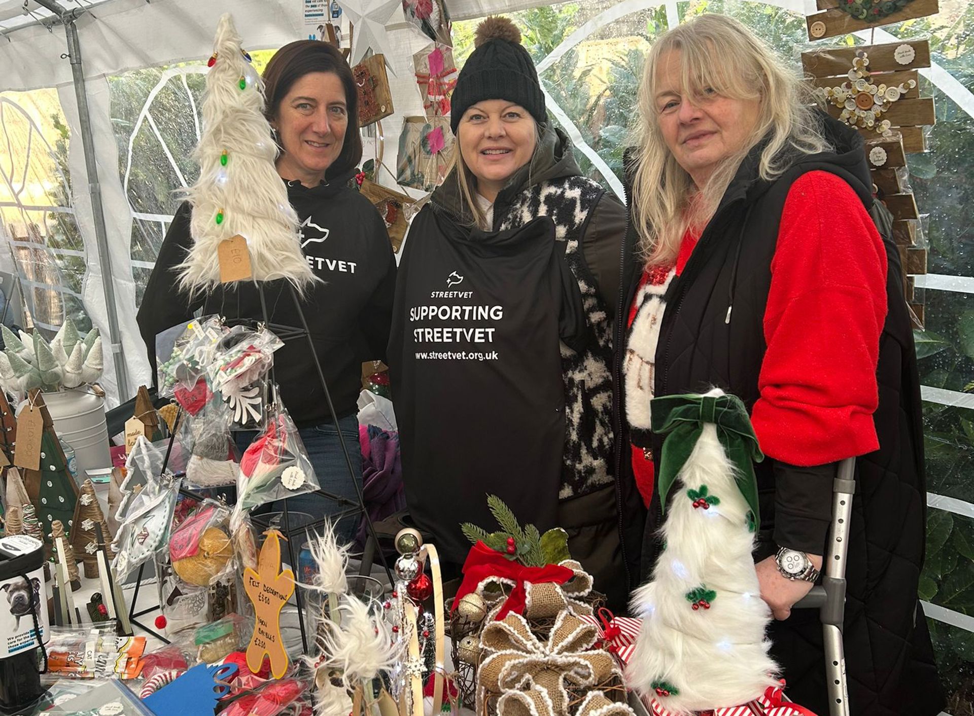 Three people stand behind a busy market stall under a white canopy, surrounded by handmade crafts including wooden decorations, dog themed ornaments, wrapped gift bundles, and a small artificial Christmas tree. The stall displays a variety of items laid out on tables and hanging from the frame, with greenery visible outside the tent.