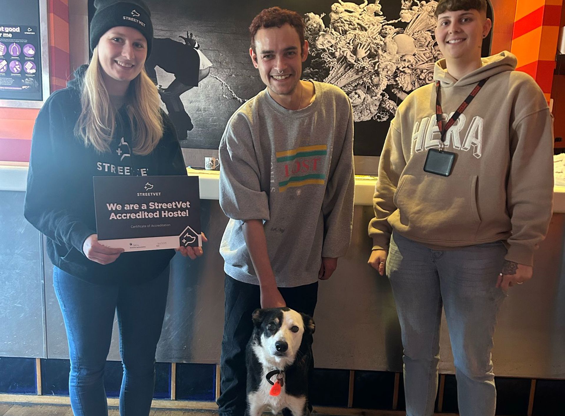 Three people, one in a StreetVet uniform is holding a certificate stand with a black-and-white dog inside at a StreetVet Accredited Hostel. 