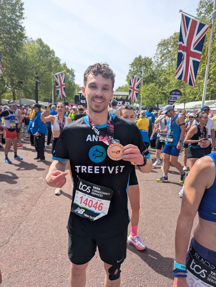 A person wearing a black running outfit with “Team StreetVet” branding and a race bib numbered 14046 stands on a wide park path among a large crowd of runners. The person holds up a finisher medal on a ribbon around the neck. Several other participants in running gear are visible in the background, along with multiple Union Jack flags lining the route and trees on either side. 