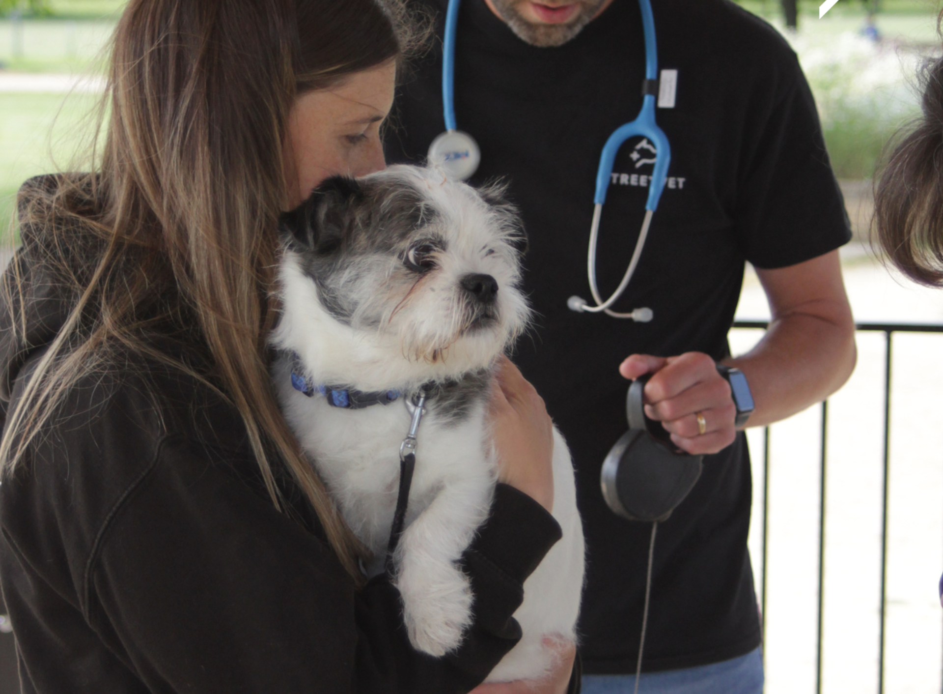 A person holds a small white and grey dog wearing a collar during an outdoor veterinary check. Another person wearing a black StreetVet T shirt with a stethoscope around the neck stands nearby, preparing or examining equipment. The setting appears to be a covered outdoor area with a railing and greenery visible in the background.